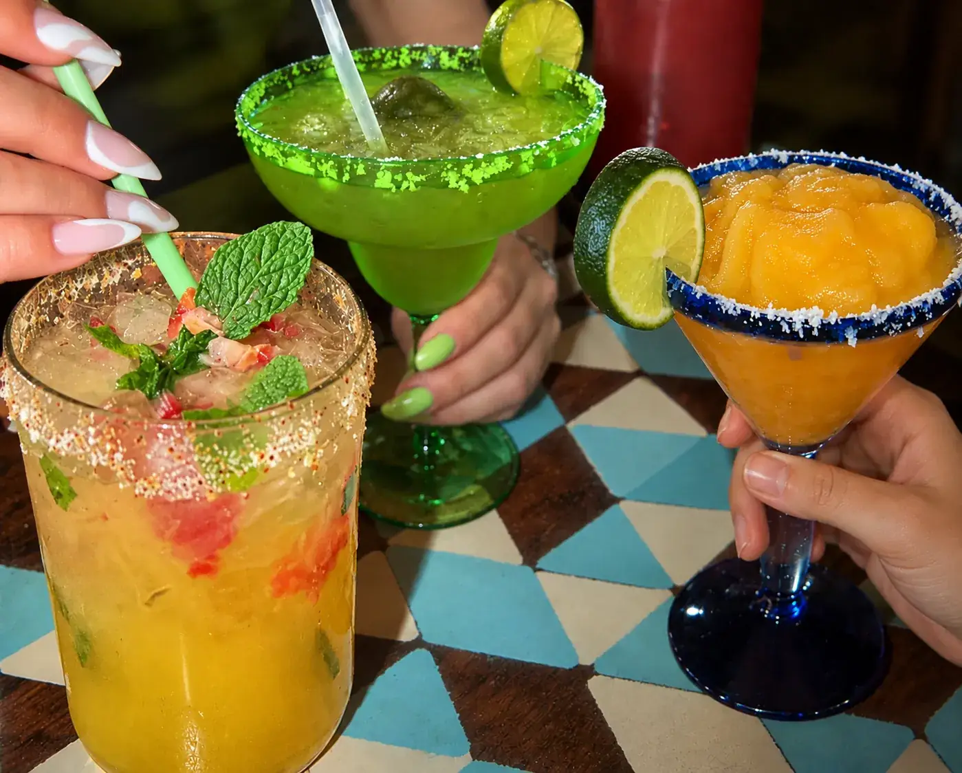 Three colorful cocktails garnished with fruit, raised together for a toast